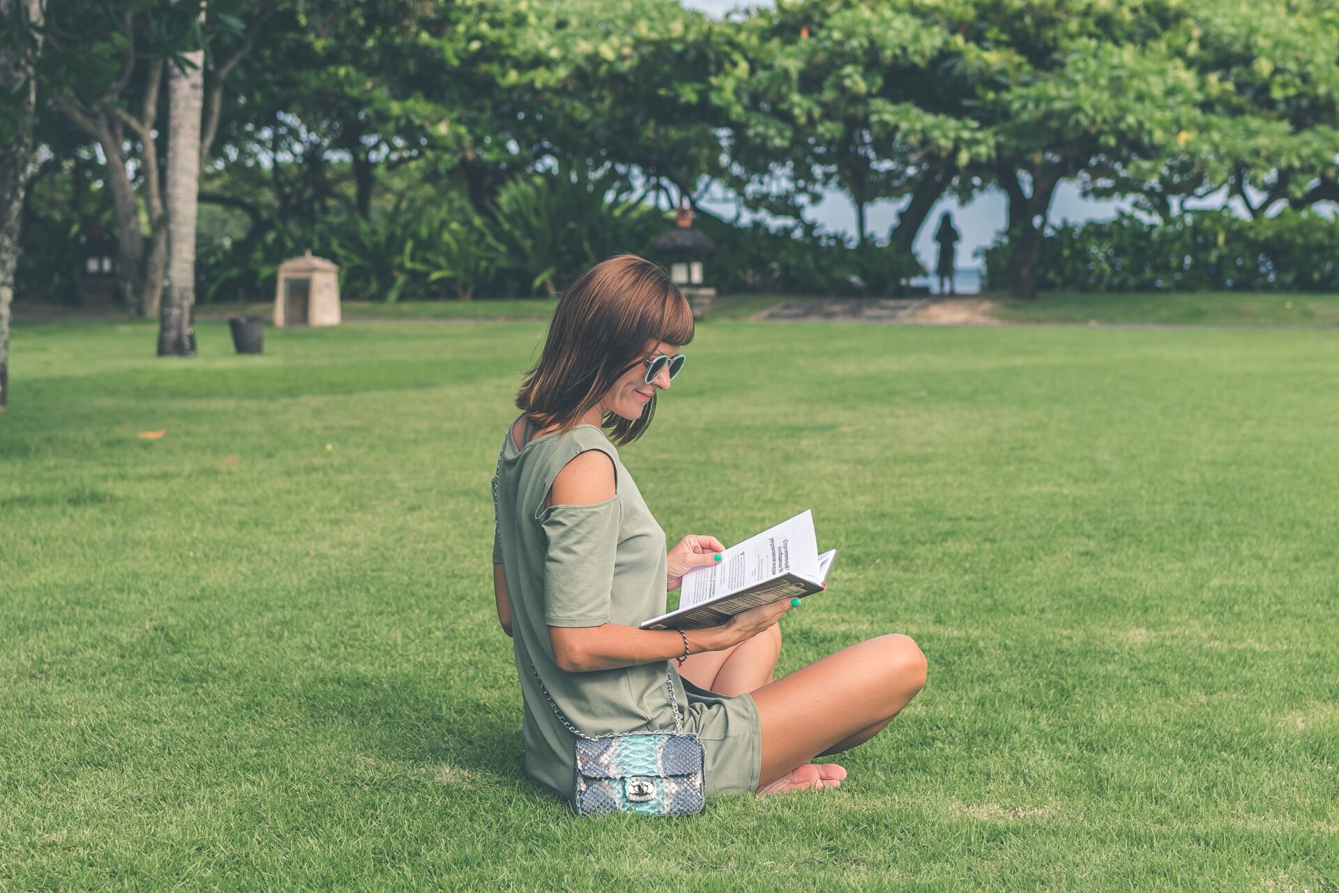 girl in blue shorts and grey t-shirt, sits on green grass in a park reading a book