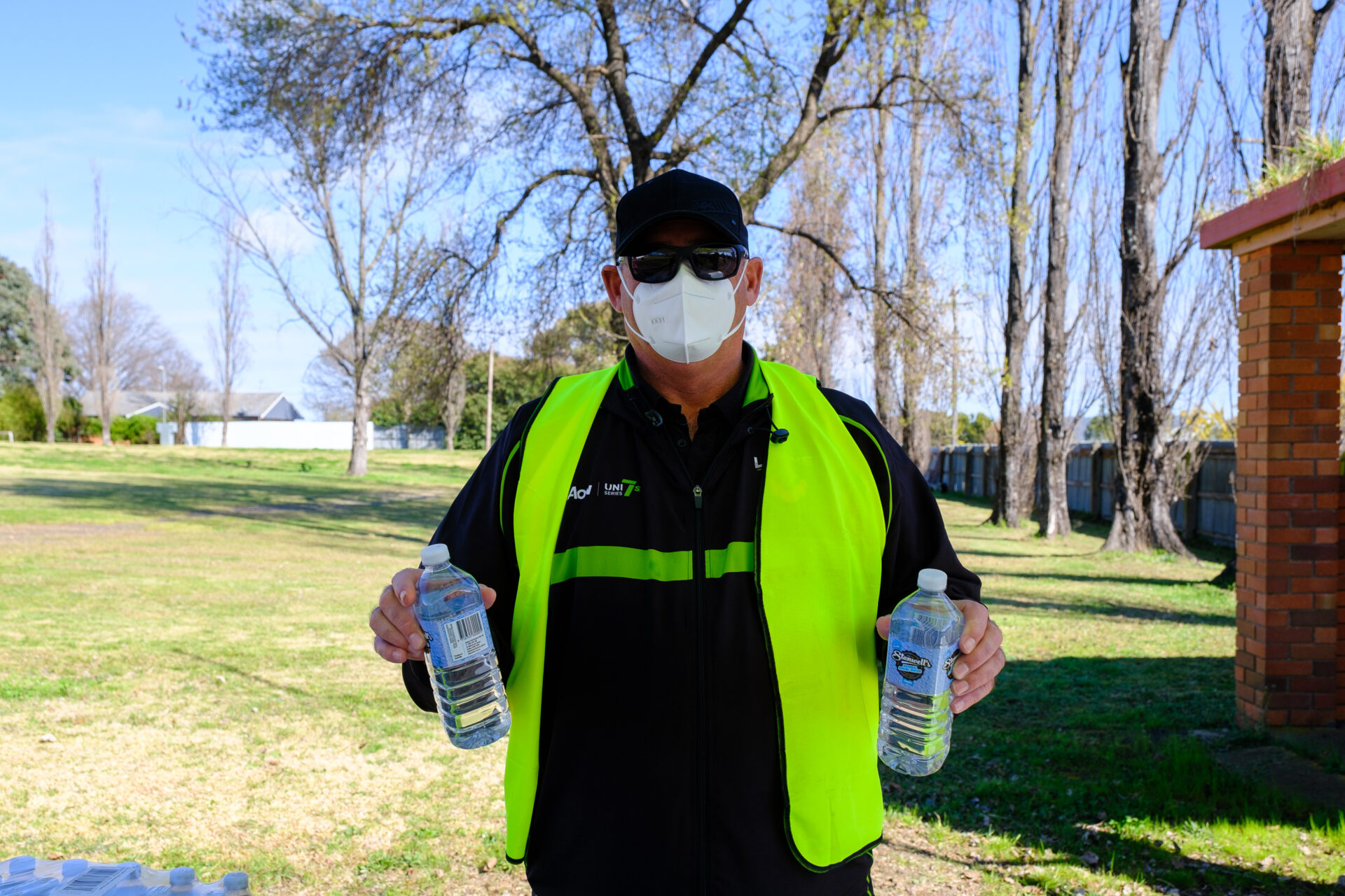 Alan stand holding two water bottles and smiling at the camera whilst wearing a high-vis vest and mask